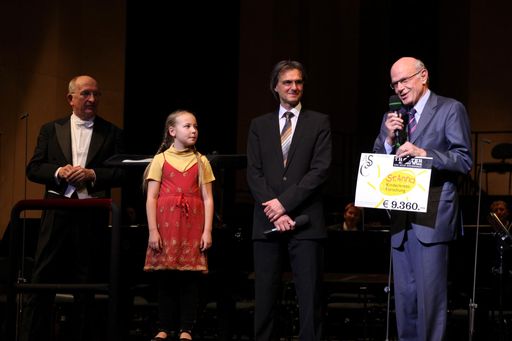 Vorschau Bild von Intendant Roland Geyer und Kinderchoristin Maria von den Gumpoldskirchner Spatzen überreichen Prof. Dr. Helmut Gadner einen Spendenscheck über 9.360,- Euro auf der Bühne des Theater an der Wien.
