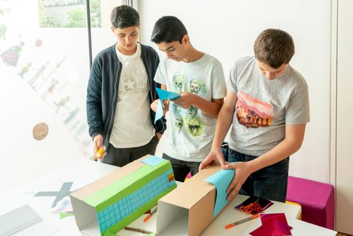 Vorschau Bild von Workshop-TeilnehmerInnen bauen am Modell weiter,
mit eigenen Gebäuden aus Papier und Karton.