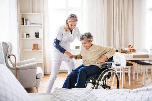 Vorschau Bild von A young health visitor helping a senior woman to
stand up from a wheelchair at home.