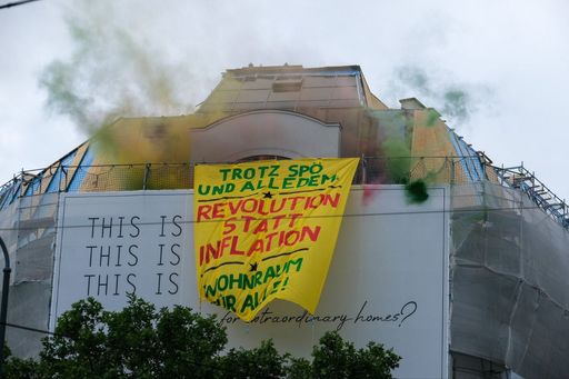 Ein Banner hängt von einem Gerüst, das zum Bau
teurer Dachgeschosswohnungen an einem Gebäude angebracht ist. Auf
dem Banner steht : "Trotz SPÖ, trotz alledem: Revolution statt
Inflation - Wohnraum für alle!". Es ist der Rauch
verschiedenfarbiger Nebelkerzen zu sehen.