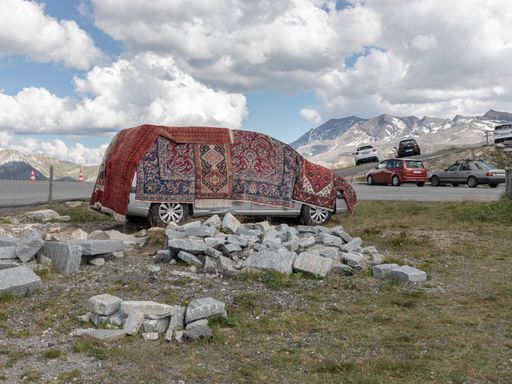 Vorschau Bild von Über und über mit Knüpf- und Webteppichen behängt
verweist NOMAD auf die mühsamen Alpenüberquerungen entlang
historischer Güterwege, auf den Transport von Kulturgütern, ebenso
aber auch auf liebevoll beladene Familienautos, die das Hab und Gut
aus Ländern des globalen Südens nach Norden tragen.