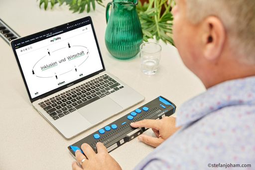  Eine Person sitzt vor dem Laptop und bedient diesen mit
Braille-Zeile. Auf dem Bildschirm ist die Webseite www.myability.org
geöffnet. Dort steht geschrieben: Inklusion und Wirtschaft.