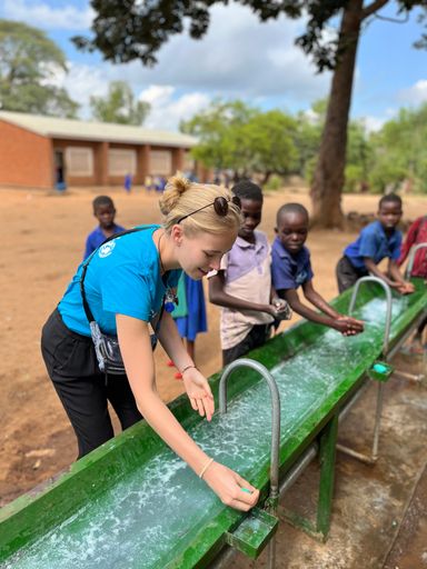 Vorschau Bild von Valerie Huber bei dem Programmbesuch in Malawi. Sie
wäschts sich neben Schüler:innen die Hände, die Schule und das
umliegende Dorf haben dank der solarbetriebenen Wasserpumpen jetzt
einen beständigen Zugang zu sauberem Wasser.