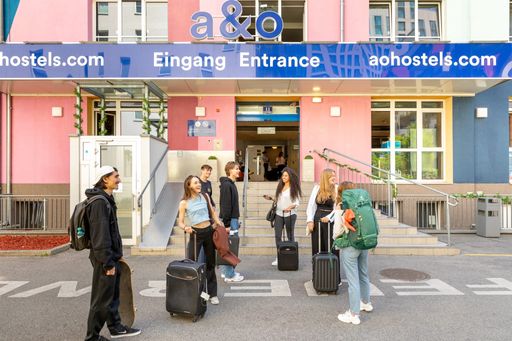 Eine Gruppe junger Leute mit Gepäck, die vor dem
a&o Hostel am Wiener Hauptbahnhof stehen. Im Hintergrund sind die
Eingangstür und das a&o-Firmenlogo zu sehen. Die Fassade des
Hostelgebäudes ist Rosa und Blau gefärbt.