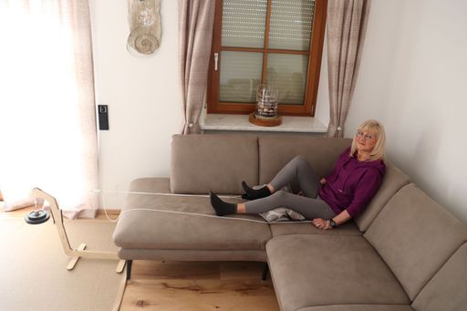 Vorschau Bild von Der flextrainer besteht aus hochwertigem
Birkensperrholz, einer Fußmanschette, Klemme, Seilen, und
Hantelgewichten. Das Bild zeigt die Anwendung am rechten Bein (Knie-
und Hüftgelenk), vorzugsweise wird im Wohnzimmer etwa beim Fernsehen
oder Lesen trainiert. Nach dem Anlegen der Fußmanschette sollte der
Zug bis zu einer Stunde gehalten werden, was in der Regel als sehr
angenehm empfunden wird. Für einen guten Erfolg sollte die Therapie
mehrmals wöchentlich durchgeführt werden.