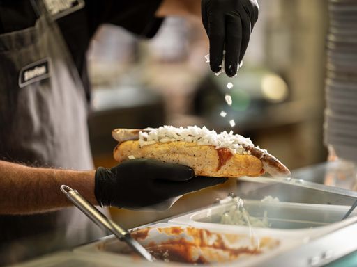 Vorschau Bild von Jürgen Ecker, Profi Koch und Mitbegründer von Bosna Plus, streut
frisch gehackten weißen Zwiebel über die frisch gegrillte Bratwurst
im eigenen Bosna Brot.