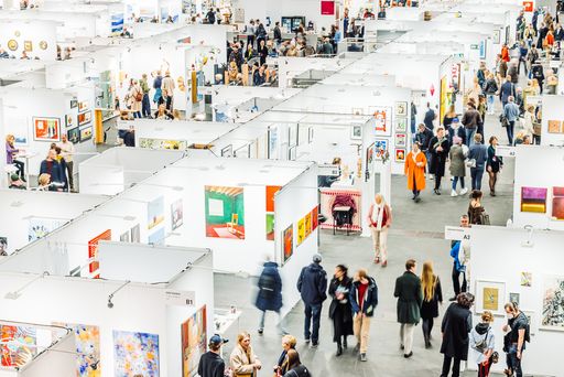  Eine Kunstmesse mit weißen Ausstellungsständen, bunten
Kunstwerken und vielen Besuchern.
