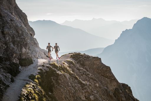  Zwei TrailrunnerInnen laufen über eine Bergkuppe hinauf.