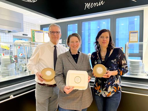  Ein Gruppenbild mit drei Personen vor einer Restauranttheke. Jede
Person hält jeweils eine braune Einwegbox mit einem "Mittagessen mit
Sinn"-Pickerl auf dem Deckel in den Händen.