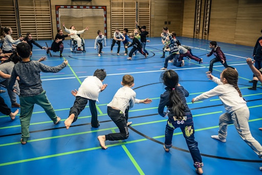  Viele Kinder und zwei erwachsene Künstler*innen sind in einem
Kreis und laufen freudig aufgeregt auf die Mitte zu. Arme fliegen
dabei in die Luft. Eine der erwachsenen Personen nutzt einen
Rollstuhl.