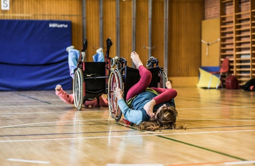 Vorschau Bild von Künstlerin Vera Rosner bei einer Performance – rücklings im
Rollstuhl, in einer ausdrucksstarken Pose. Kleidung in Blau, Grün
und Weinrot.