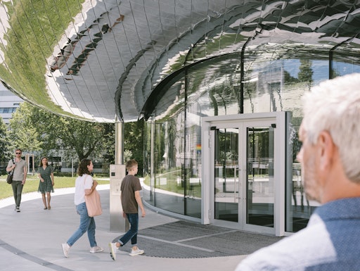  Eingang zum VISTA Science Experience Center. Die Glasfassade und
das verspiegelte Dach fangen das üppige Grün des Parks und die
angrenzenden historischen und modernen Gebäude des ISTA Campus ein
und reflektieren sie. Durch die Spiegelung der Umgebung fällt das
Gebäude ins Auge und verschmilzt gleichzeitig mit der Landschaft.