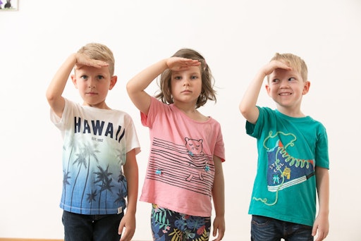 Drei Kinder stehen nebeneinander und blicken mit der Hand über
den Augen in die Ferne – als Zeichen für Aufmerksamkeit und
Sichtbarkeit im Straßenverkehr, auch bei Dunkelheit.