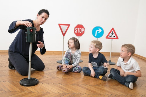 Vorschau Bild von Eine Verkehrstrainerin sitzt am Boden und erklärt drei
Kindergartenkindern spielerisch die Bedeutung der Ampel mithilfe
einer kindgerechten Spielampel.