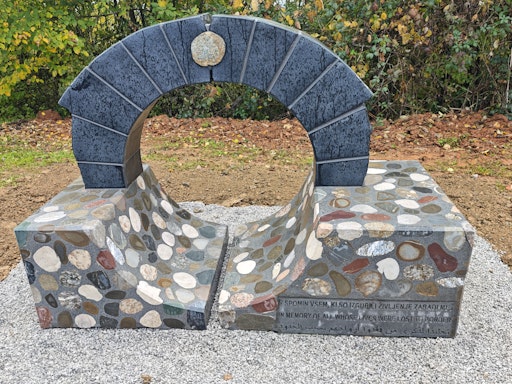 Vorschau Bild von Das Werk des akademischen Bildhauers Tomaž Furlan hat die Form
einer Brücke bzw. eines Übergangs. Der Sockel besteht aus Steinen
aus dem Fluss Kolpa – so sollen die Orte der Tragödien symbolisch in
das Denkmal selbst einfließen.