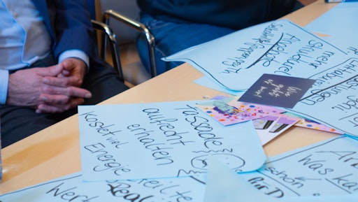 Auf einem Tisch liegen große blaue Post-Its. Auf einem kann man
lesen: Fassade aufrecht erhalten kostet viel Energie