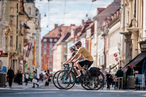 Vorschau Bild von Zwei Personen fahren mit Fahrrädern durch die belebte Grazer
Altstadt mit historischen Gebäuden und Fußgängern