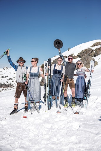 Personen in Tracht und auf Ski stehen gut gelaunt am Pistenrand