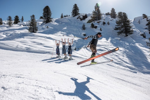 Vorschau Bild von Einer in Lederhose springt mit Ski über eine Pistenkante, Mädels
im Dirndl jubeln ihm zu