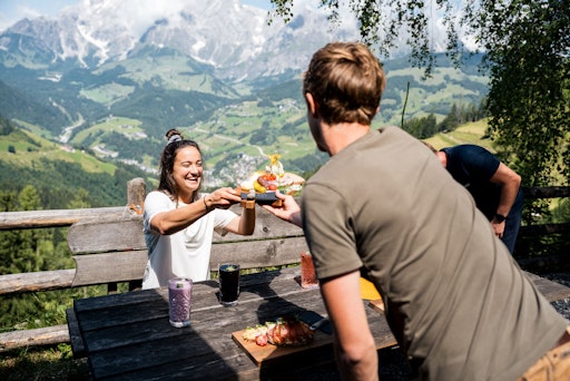 Frau nimmt ihre Kräuterjause lachend vom Kellner entgegen, im
Hintergrund ist das Hochkönig-Panorama zu sehen.