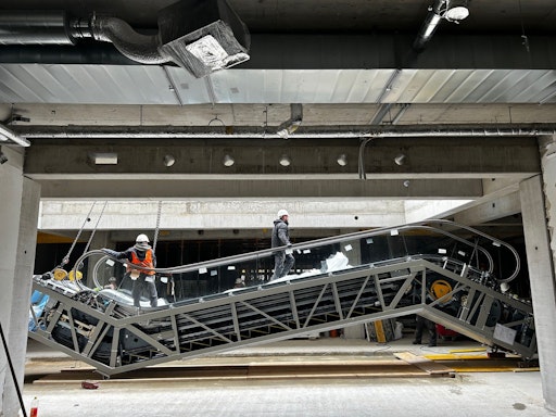 Vorschau Bild von 2 Monteure gehen über eine am Boden stehende Fahrtreppe, die über
Stahlseile in das Gebäude eingebracht wurde.