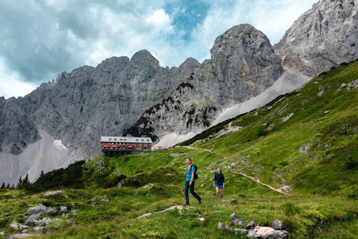 Zwei Personen sind auf der Sternwanderung "Kaiserblüte" am Wilden
Kaiser unterwegs, im Hintergrund die Berge sowie Hütte zu sehen.
