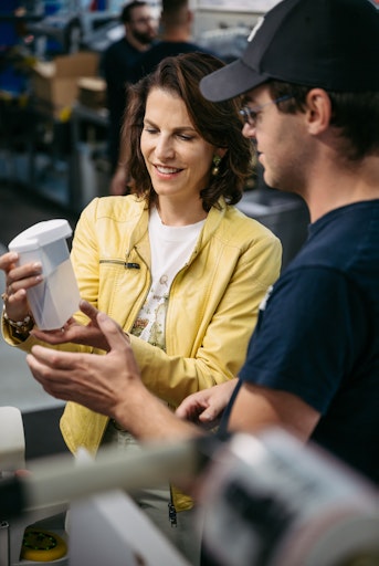 Vorschau Bild von Karoline Edtstadler mustert einen fertigen Hygienespender –
gemeinsam mit Stefan Fuchslechner, der an der Produktionslinie
mitwirkt (im Bild von links)