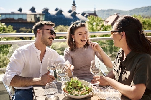 Vorschau Bild von Drei Personen essen und trinken auf einer Terrasse, im
Hintergrund sind das Kunsthaus Graz und Kirchtürme zu sehen.