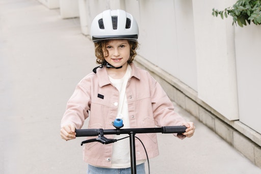 Ein junges Mädchen steht mit ihrem Roller auf einem Gehweg. Sie
trägt dabei einen Schutzhelm.