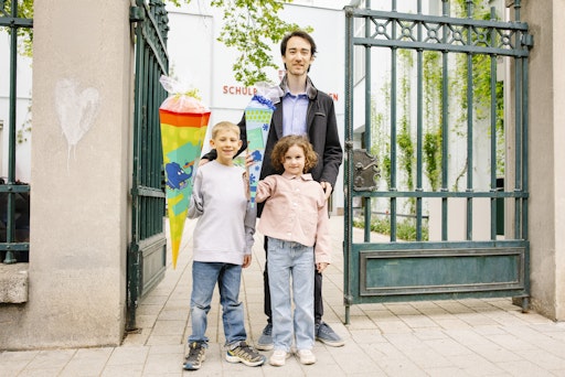 Vorschau Bild von Ein Erwachsener steht mit zwei Kindern vor einem Schultor. Der
Junge und das Mädchen halten jeweils eine Schultüte in der Hand und
lächeln in die Kamera.