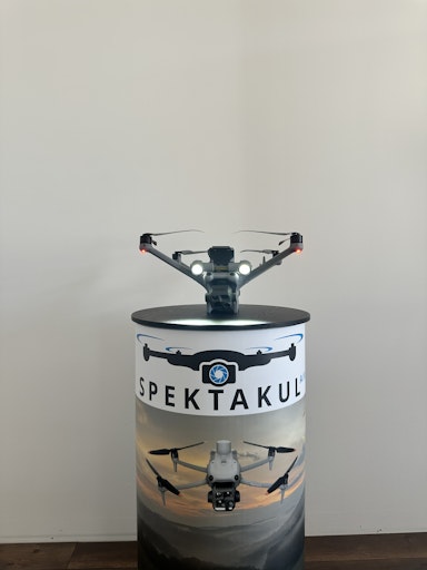 Vorschau Bild von Eine DJI Mavic 3 Enterprise Drohne, bereit für den Einsatz,
präsentiert auf einem Werbedisplay von spektakulAIR. Dieses Modell
wird häufig für professionelle Inspektionen, wie z.B. von
Photovoltaikanlagen mittels Thermografie, eingesetzt. Die
eingeschalteten Scheinwerfer unterstreichen die Einsatzbereitschaft.